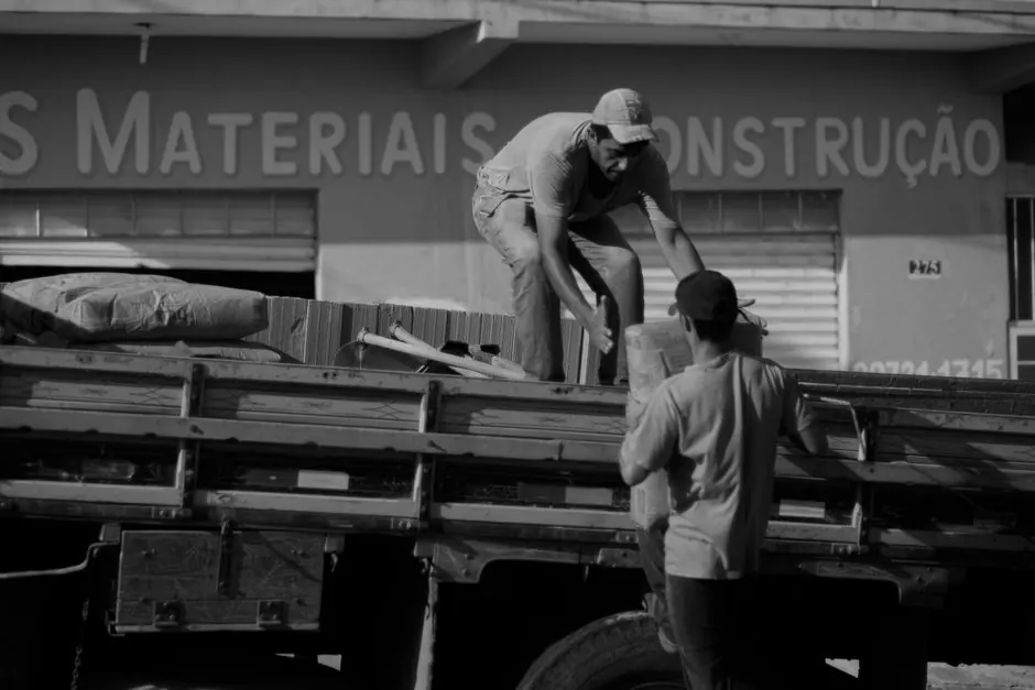 Black and white of anonymous male worker giving bag to coworker standing on trunk while working in industrial district on street